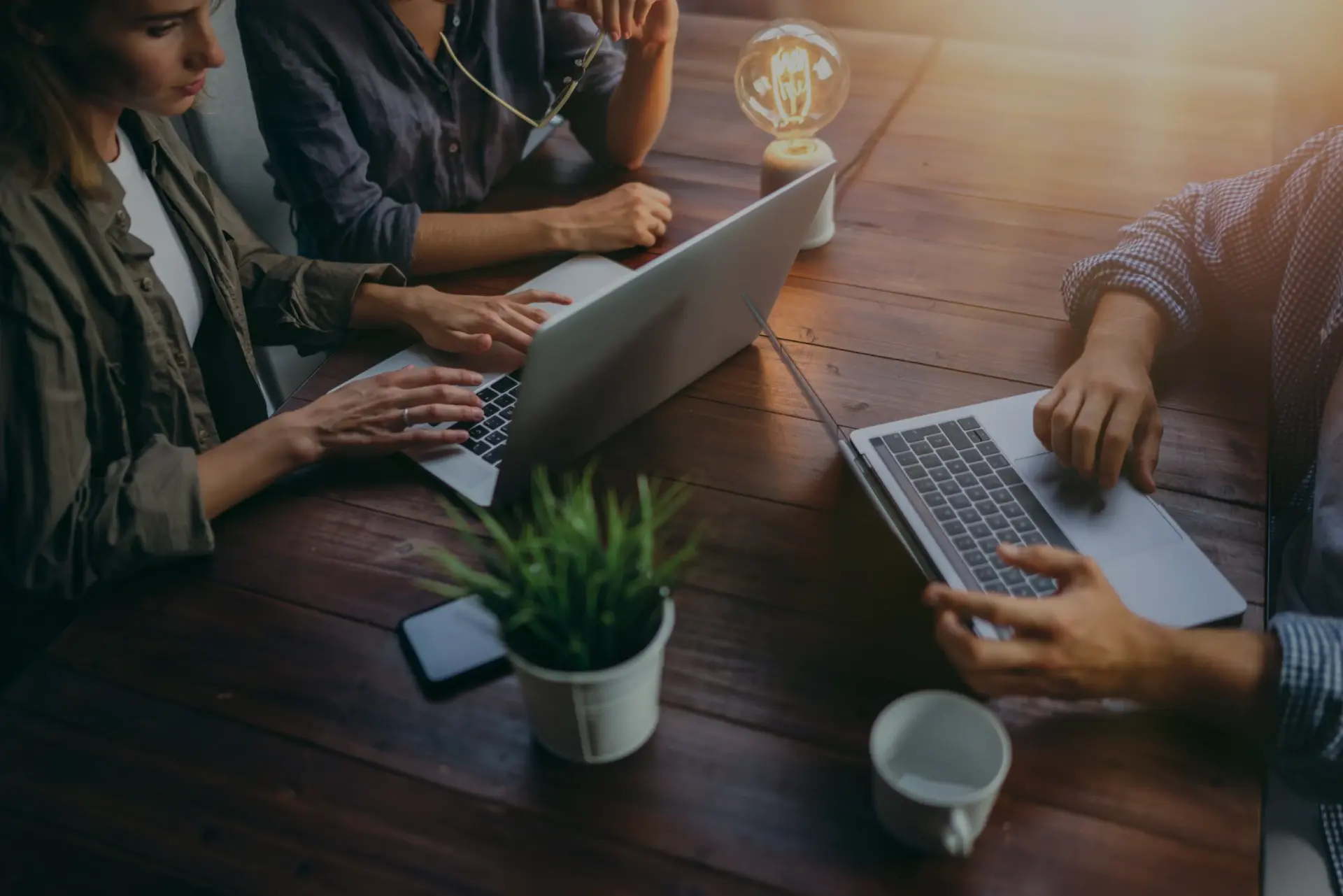 Three people are working at a wooden table with laptops and a glowing light bulb.