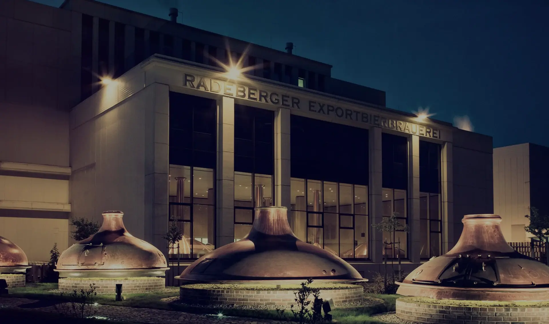 A night view of a brewery building with illuminated copper vats in front.
