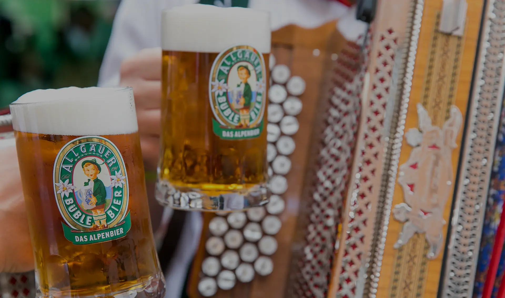 A close-up of two beer glasses filled with golden beer, foam at the top, in front of a blurry accordion.