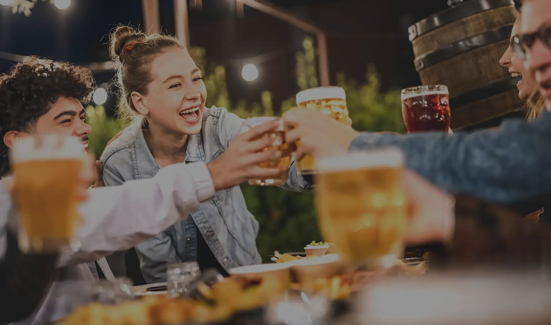 Four young adults enjoy a cheerful toast with beers at a warmly lit outdoor table.